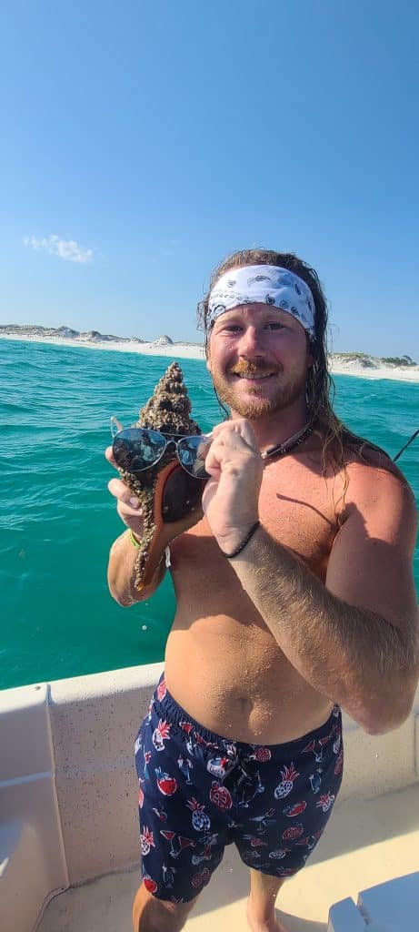 Young man holding up a conch shell with sunglasses he found on a private boat tour with Anchor Charters in Panama City Beach Florida